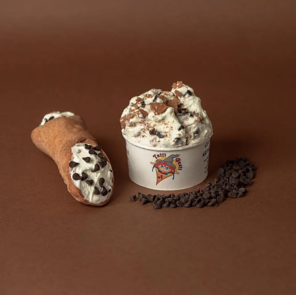 Coppetta di gelato al cannolo accanto a un cannolo siciliano e gocce di cioccolato su fondo cacao
