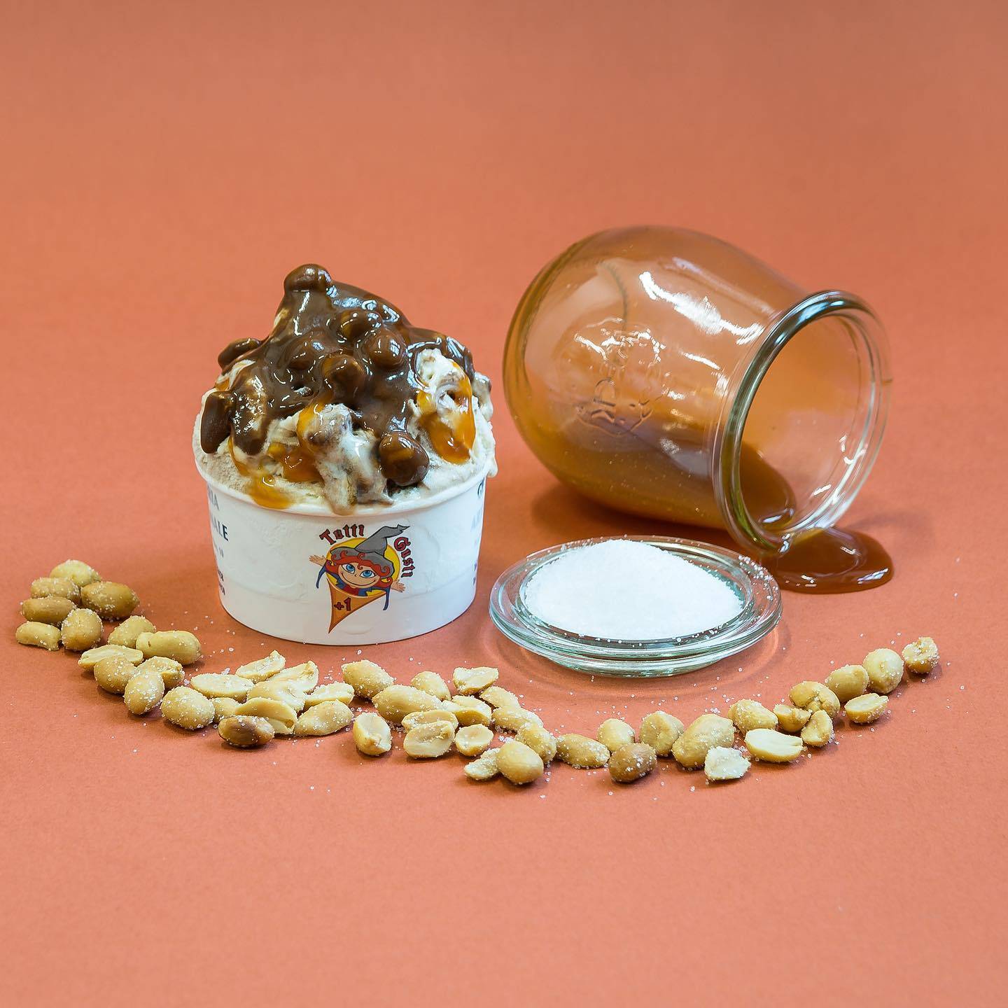 Coppetta di gelato al caramello con arachidi e un vasetto di caramello rovesciato, su fondo terracotta